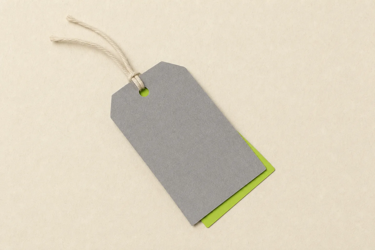 A single grey price tag lying on a bone-cream ground with a second, identical tag hidden almost entirely beneath it, only a sliver of its corner showing in solid acid green.